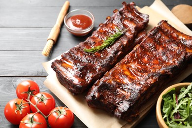 Photo of Tasty roasted pork ribs served with rosemary, tomatoes, sauce and salad on black wooden table, closeup