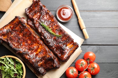 Photo of Tasty roasted pork ribs served with rosemary, tomatoes, sauce and salad on black wooden table, flat lay