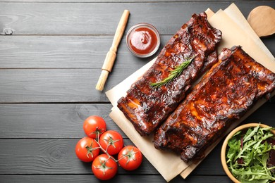 Photo of Tasty roasted pork ribs served with rosemary, tomatoes, sauce and salad on black wooden table, flat lay. Space for text