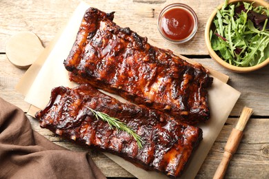 Photo of Tasty roasted pork ribs served with rosemary, sauce and salad on wooden table, flat lay