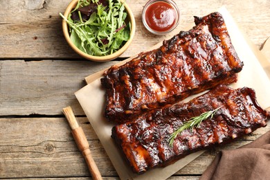 Photo of Tasty roasted pork ribs served with rosemary, sauce and salad on wooden table, flat lay