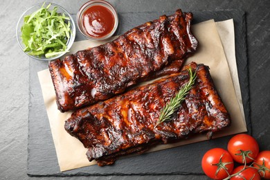 Photo of Tasty roasted pork ribs served with rosemary, tomatoes, sauce and salad on black table, flat lay