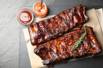 Photo of Tasty roasted pork ribs served with rosemary and sauce on black table, flat lay