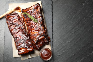 Photo of Tasty roasted pork ribs served with rosemary and sauce on black table, top view. Space for text