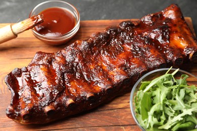 Photo of Tasty roasted pork ribs served with arugula and sauce on black table, closeup
