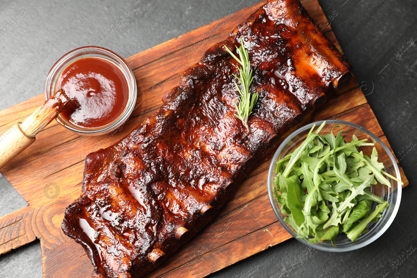 Tasty roasted pork ribs served with rosemary, arugula and sauce on black table, top view Photo of Tasty roasted pork ribs served with rosemary, arugula and sauce on black table, top view