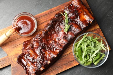 Photo of Tasty roasted pork ribs served with rosemary, arugula and sauce on black table, top view