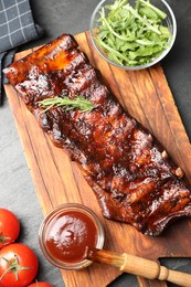 Photo of Tasty roasted pork ribs served with rosemary, arugula, tomatoes and sauce on black table, flat lay