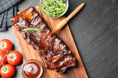 Photo of Tasty roasted pork ribs served with rosemary, arugula, tomatoes and sauce on black table, flat lay. Space for text