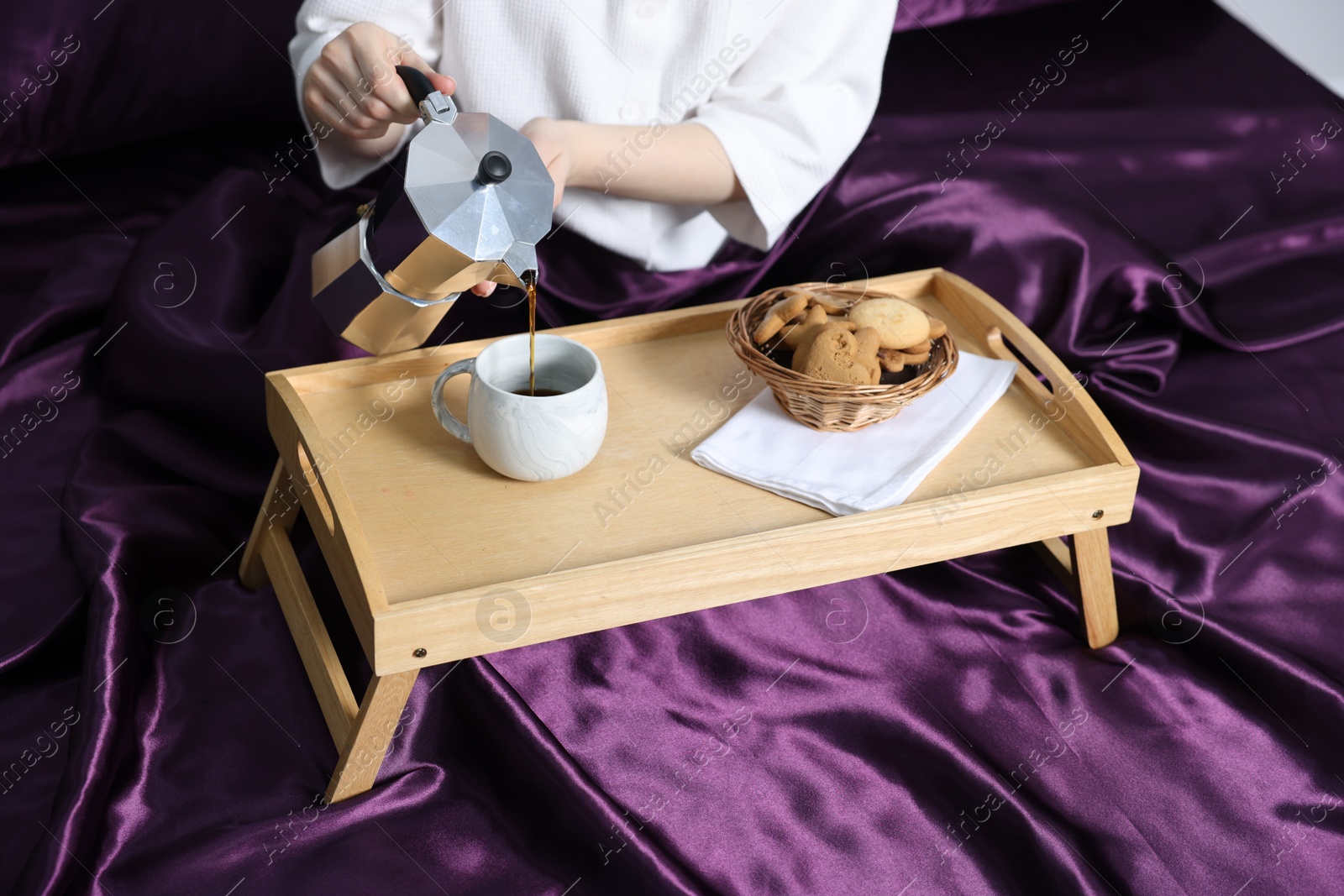 Photo of Woman pouring coffee from moka pot into cup on bed with purple silk linens, closeup