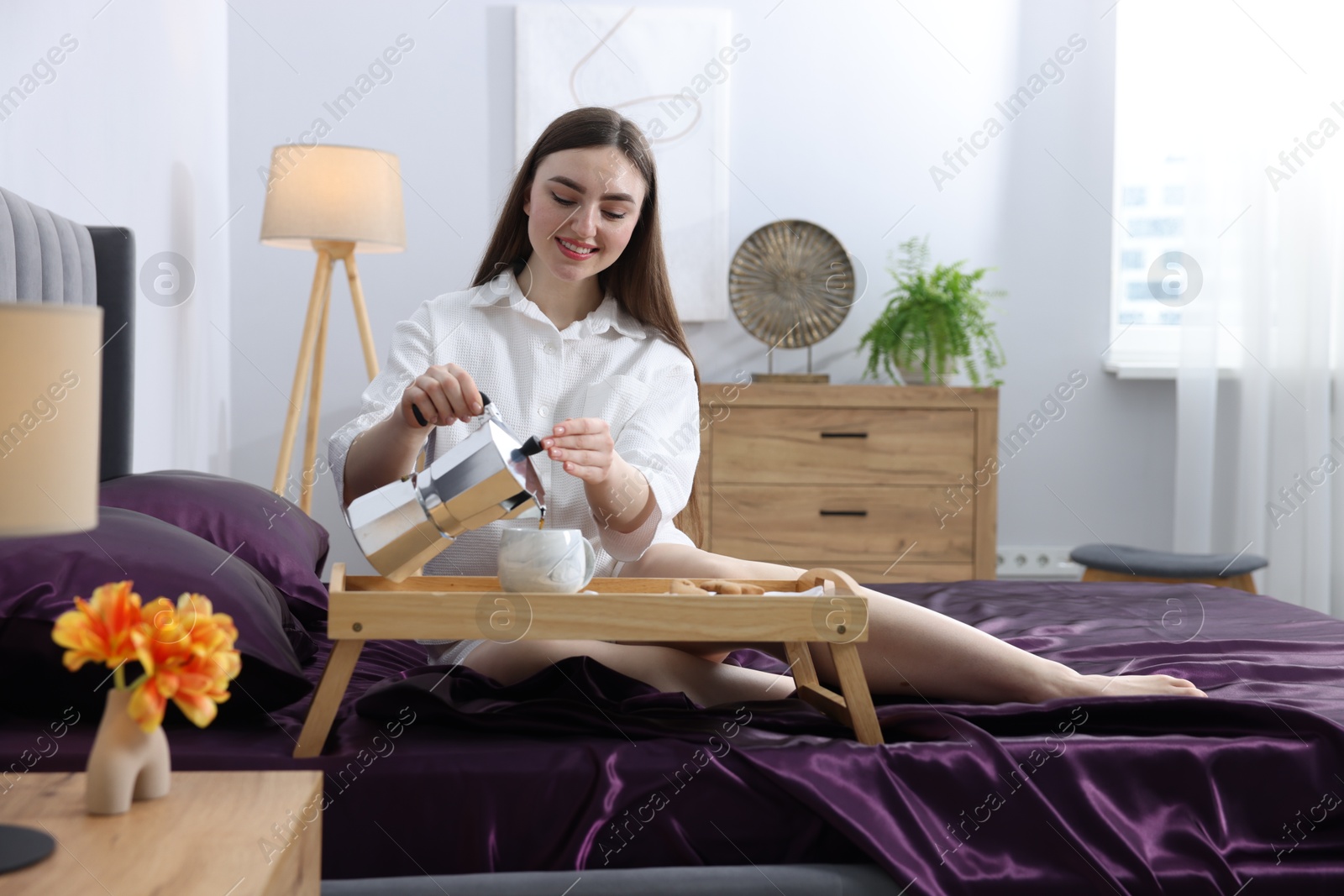 Smiling woman pouring coffee from moka pot into cup on bed with purple silk linens indoors Photo of Smiling woman pouring coffee from moka pot into cup on bed with purple silk linens indoors