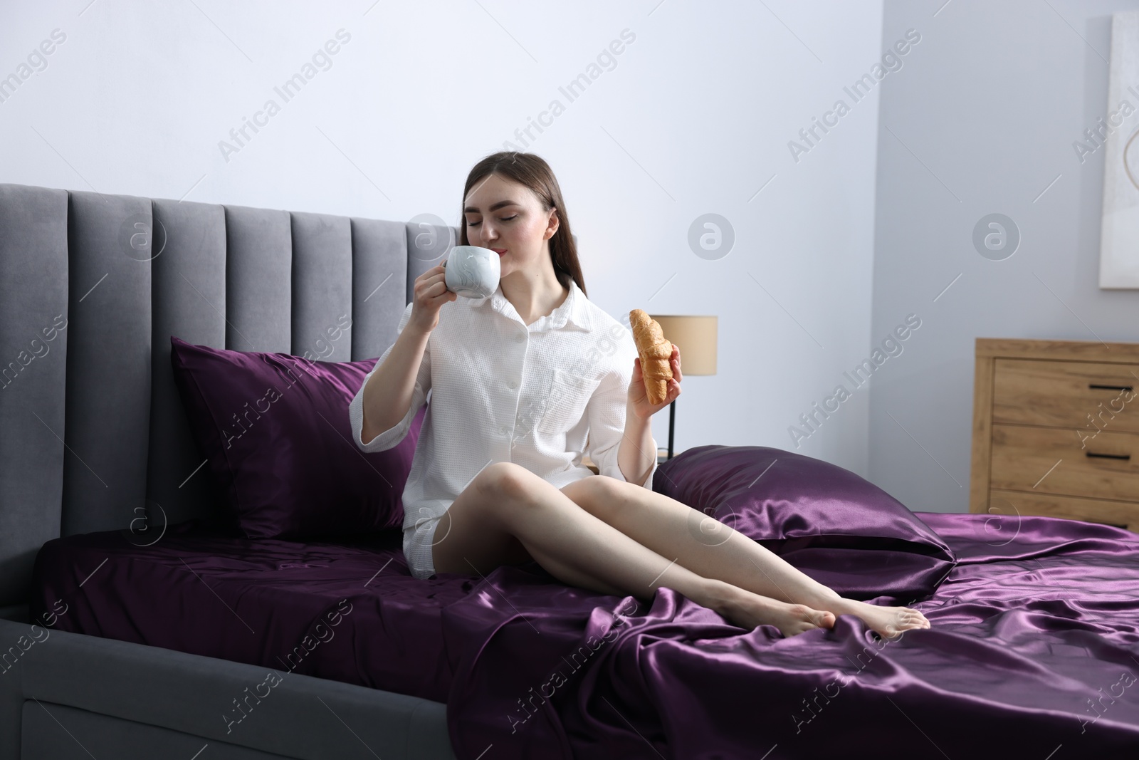Woman with coffee and croissant on bed with purple silk linens indoors Photo of Woman with coffee and croissant on bed with purple silk linens indoors