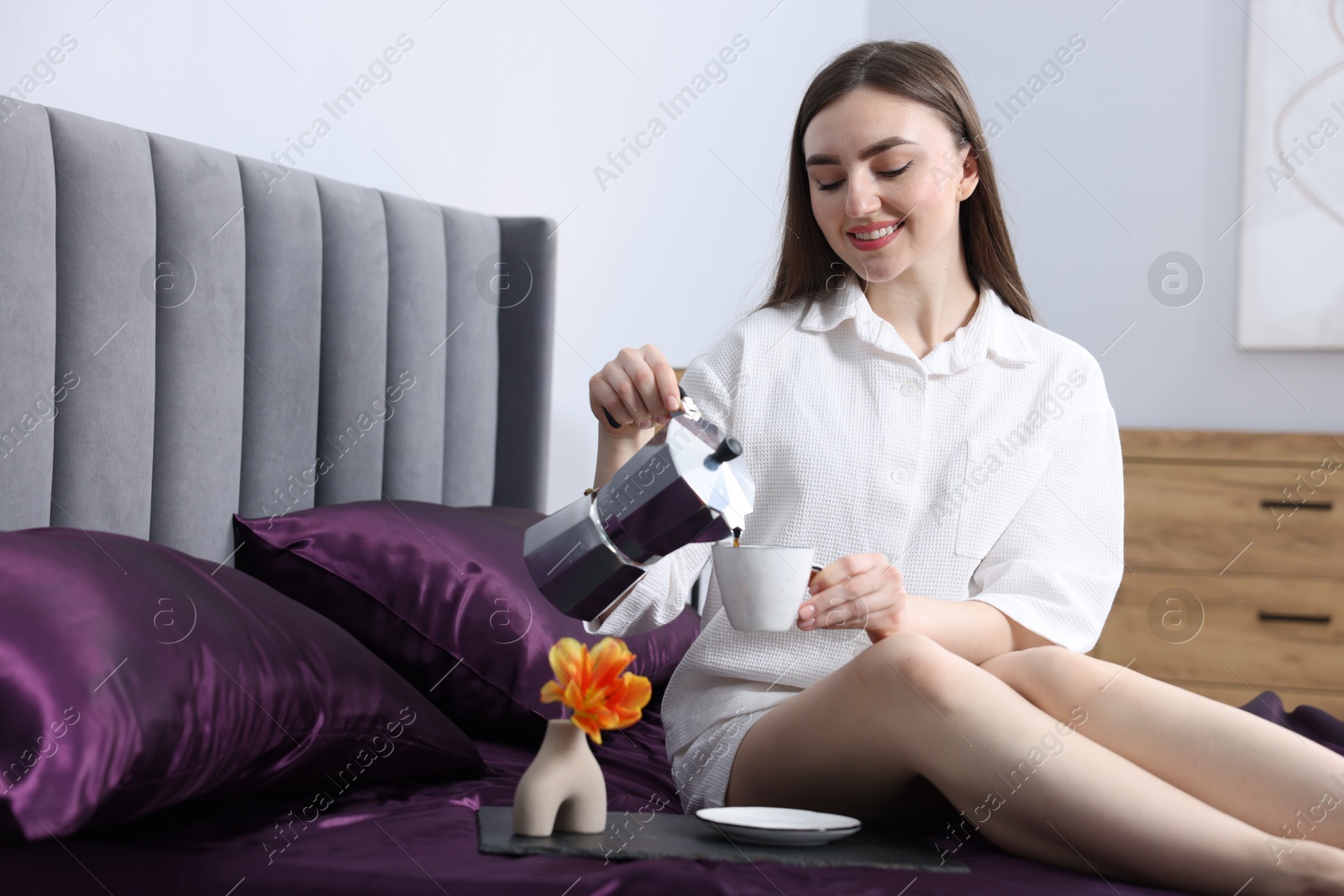 Smiling woman pouring coffee from moka pot into cup on bed with purple silk linens indoors Photo of Smiling woman pouring coffee from moka pot into cup on bed with purple silk linens indoors