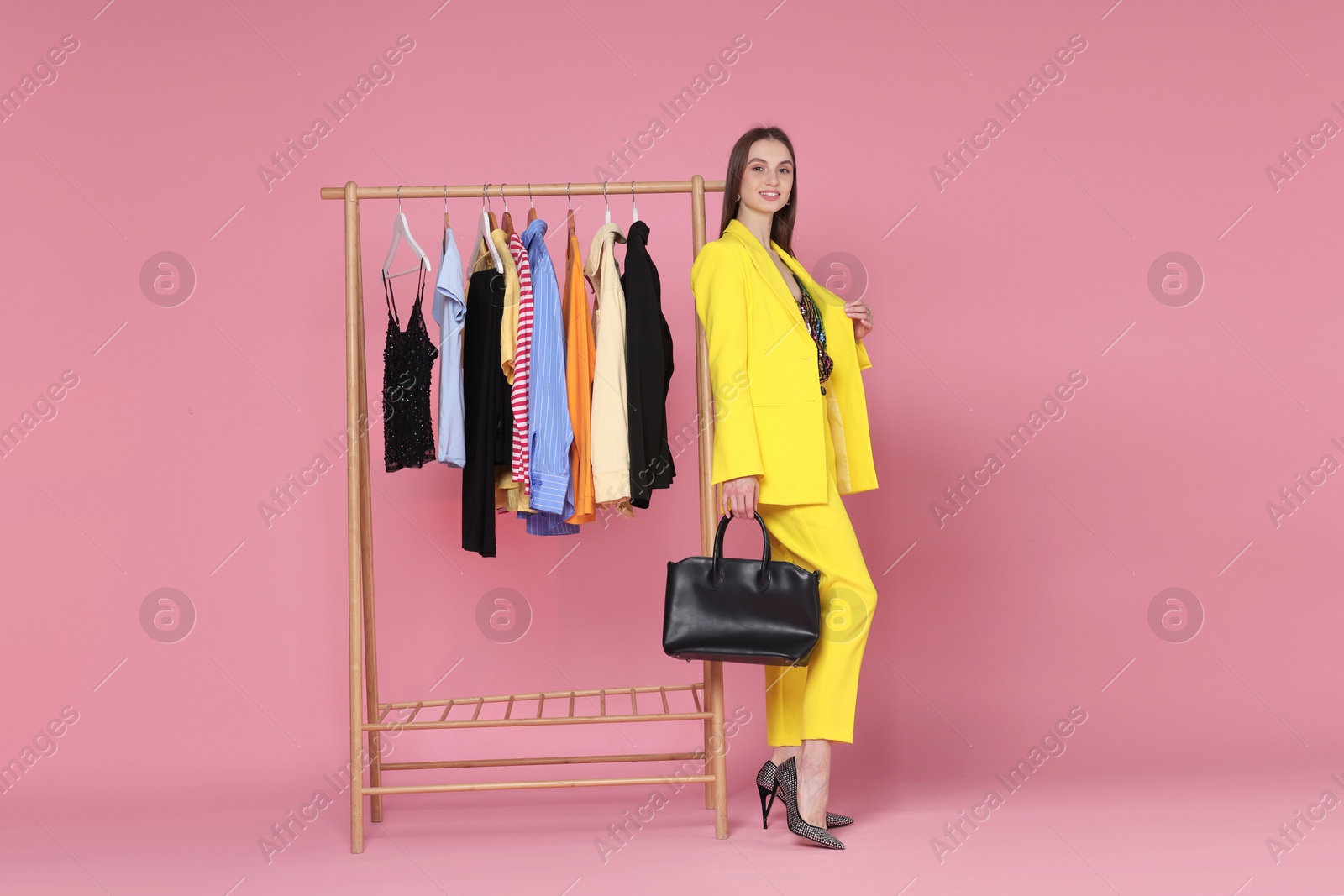 Smiling fashion stylist with bag and clothing rack on pink background Photo of Smiling fashion stylist with bag and clothing rack on pink background