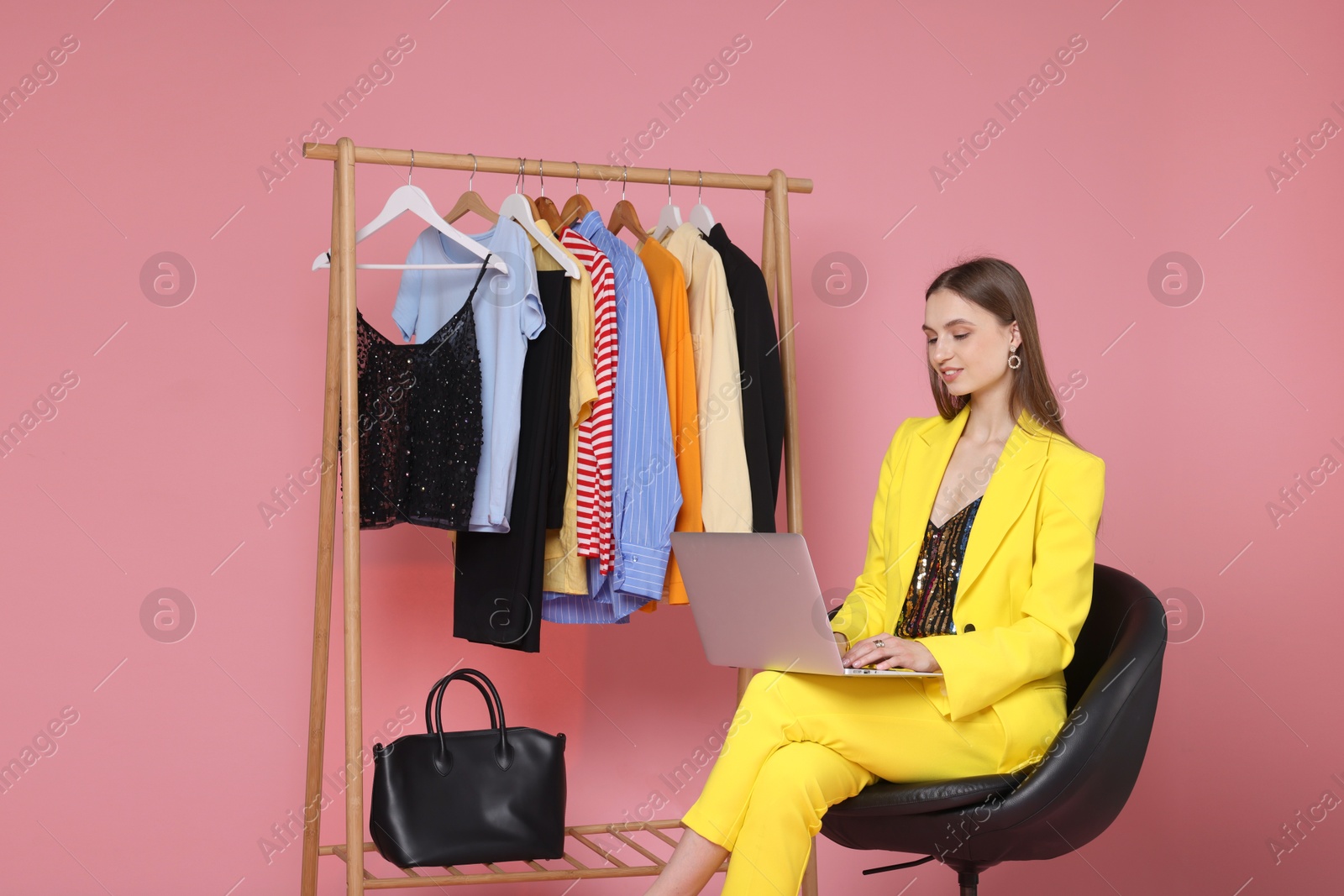 Smiling fashion stylist working with laptop and clothing rack on pink background Photo of Smiling fashion stylist working with laptop and clothing rack on pink background