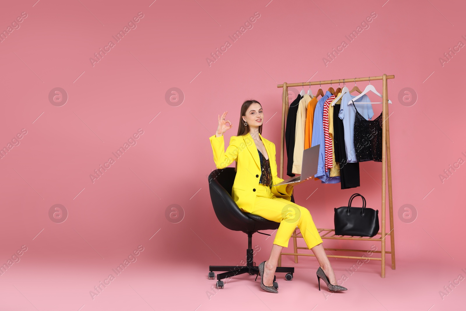 Fashion stylist with laptop and clothing rack showing ok gesture on pink background. Space for text Photo of Fashion stylist with laptop and clothing rack showing ok gesture on pink background. Space for text