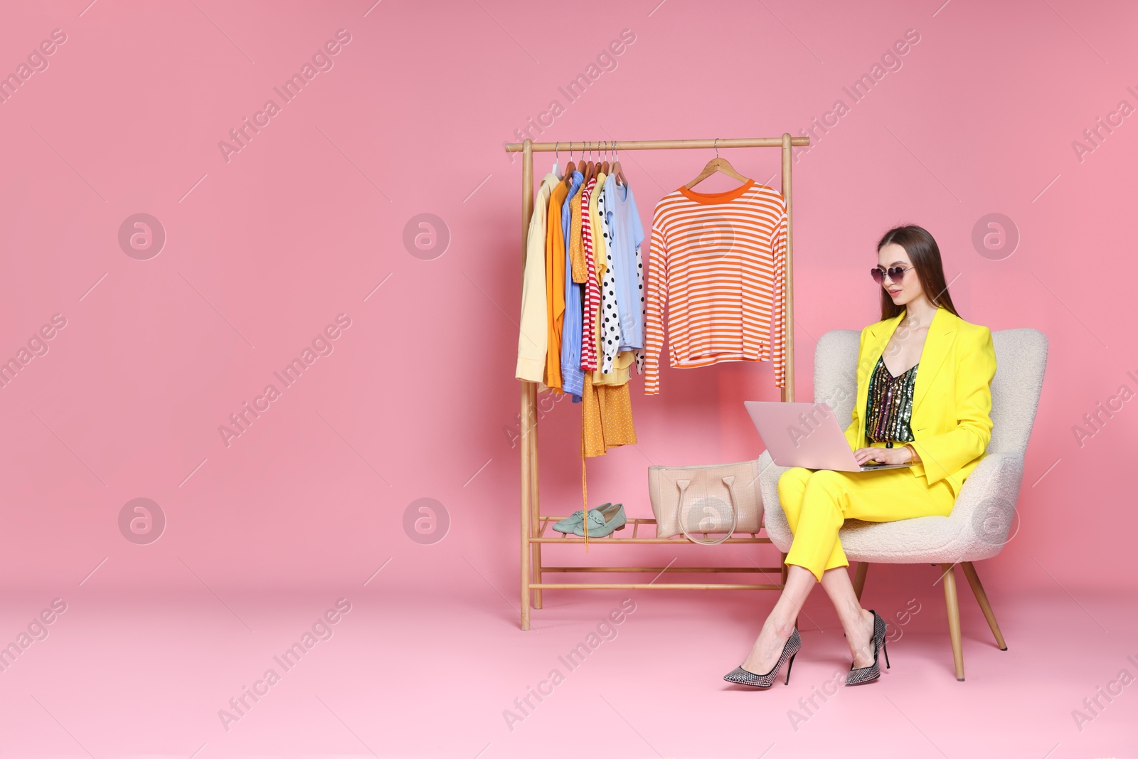 Smiling fashion stylist working with laptop and clothing rack on pink background. Space for text Photo of Smiling fashion stylist working with laptop and clothing rack on pink background. Space for text