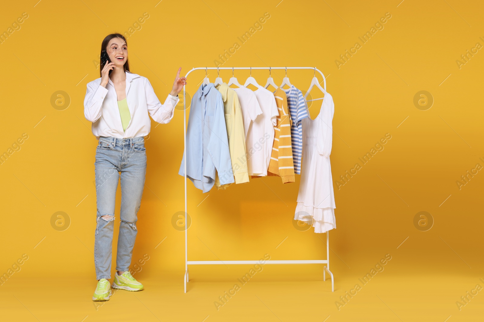 Happy fashion stylist talking by smartphone and rack with clothes on yellow background Photo of Happy fashion stylist talking by smartphone and rack with clothes on yellow background