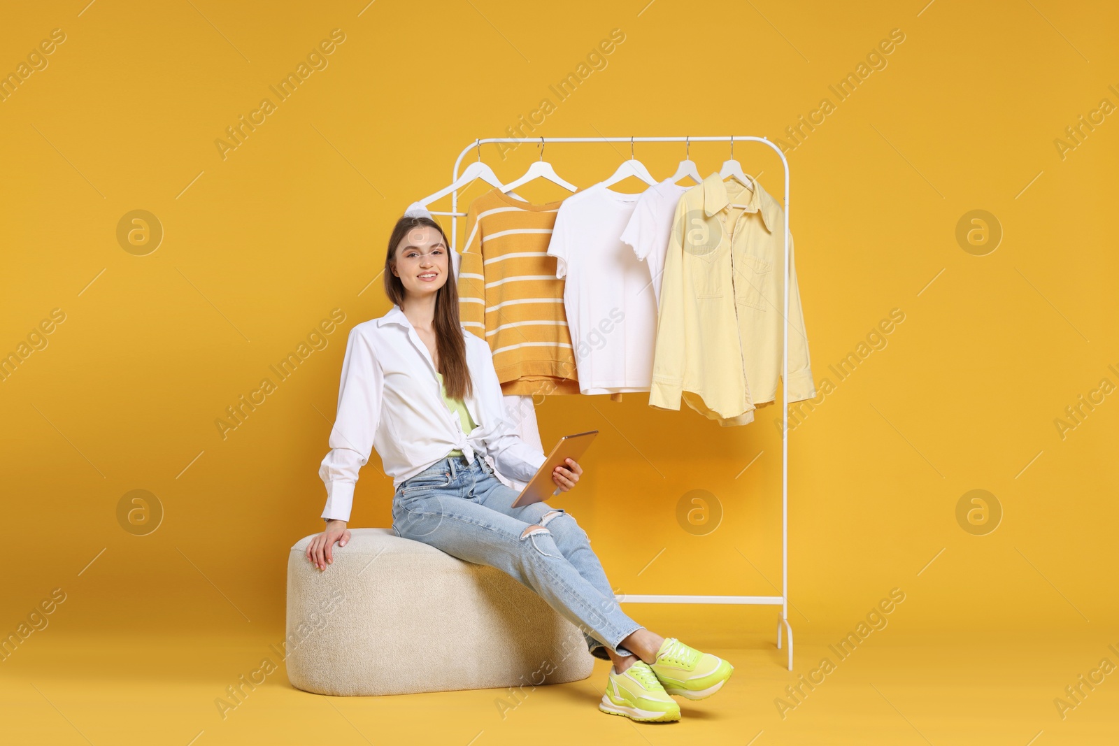 Smiling fashion stylist with tablet and clothing rack on yellow background Photo of Smiling fashion stylist with tablet and clothing rack on yellow background