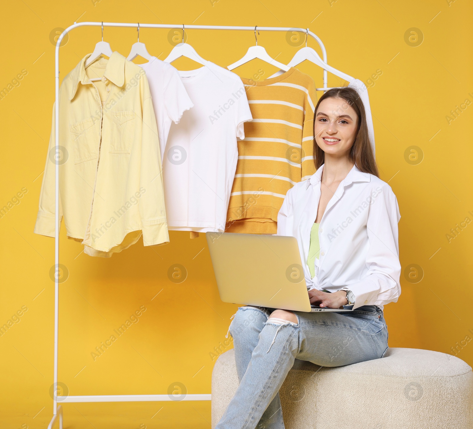 Smiling fashion stylist with laptop and clothing rack on yellow background Photo of Smiling fashion stylist with laptop and clothing rack on yellow background