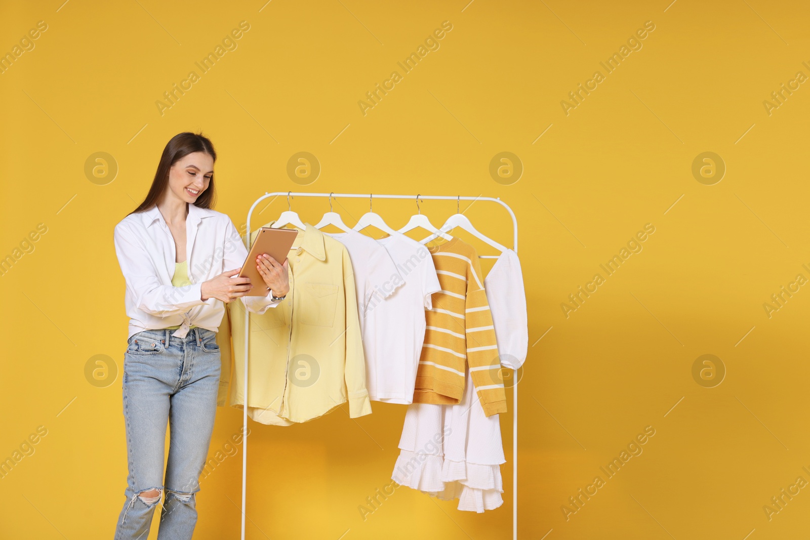 Smiling fashion stylist with tablet and clothing rack on yellow background Photo of Smiling fashion stylist with tablet and clothing rack on yellow background