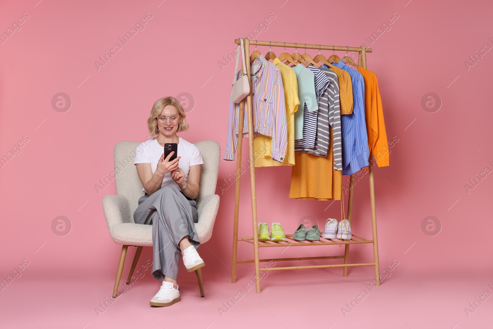 Smiling fashion stylist with smartphone and clothing rack on pink background Photo of Smiling fashion stylist with smartphone and clothing rack on pink background