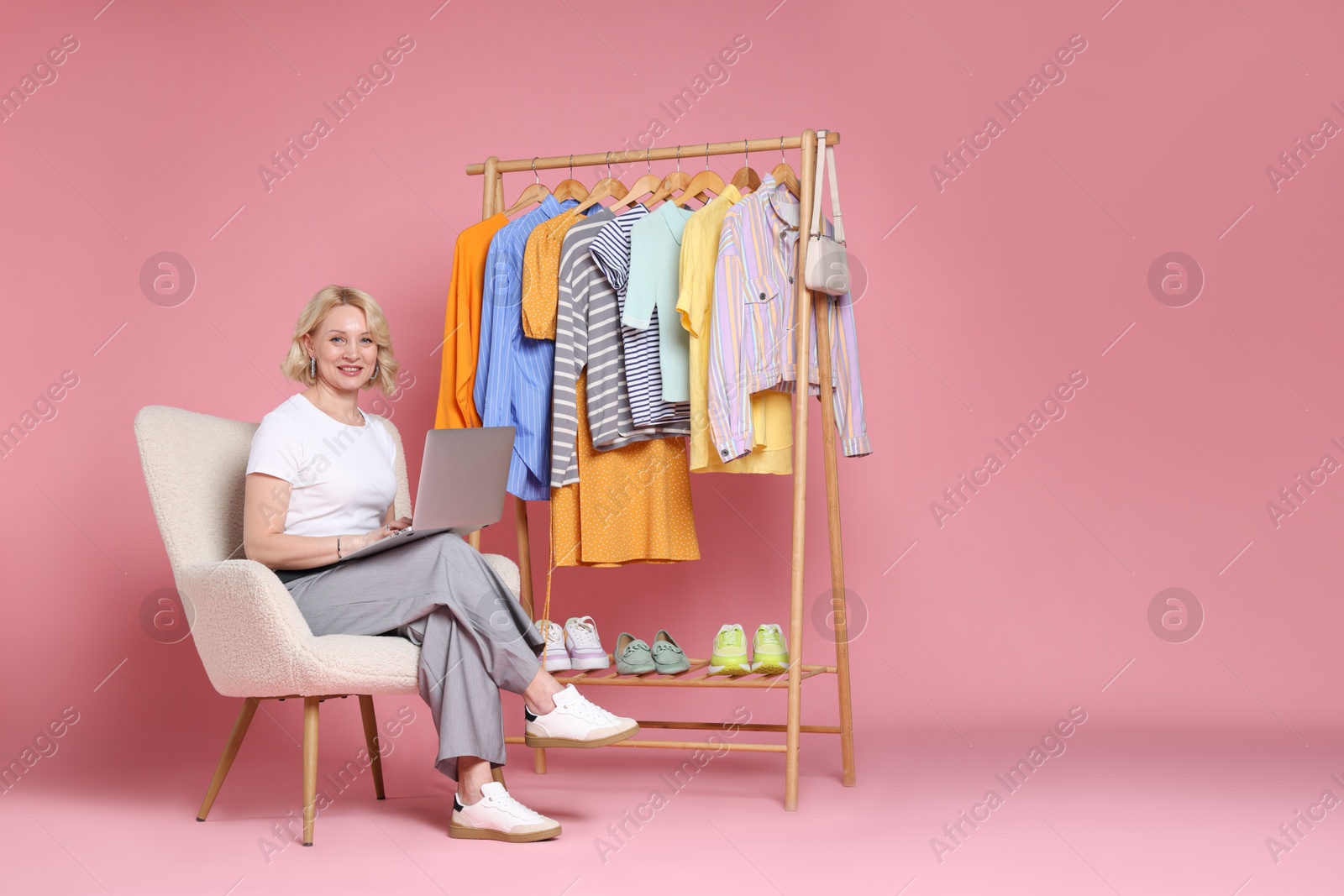 Smiling fashion stylist with laptop and clothing rack on pink background Photo of Smiling fashion stylist with laptop and clothing rack on pink background