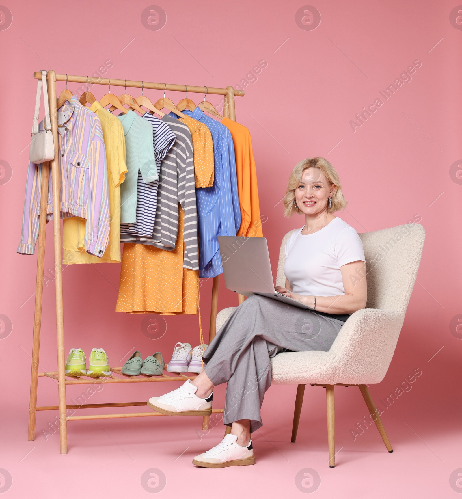 Smiling fashion stylist with laptop and clothing rack on pink background Photo of Smiling fashion stylist with laptop and clothing rack on pink background