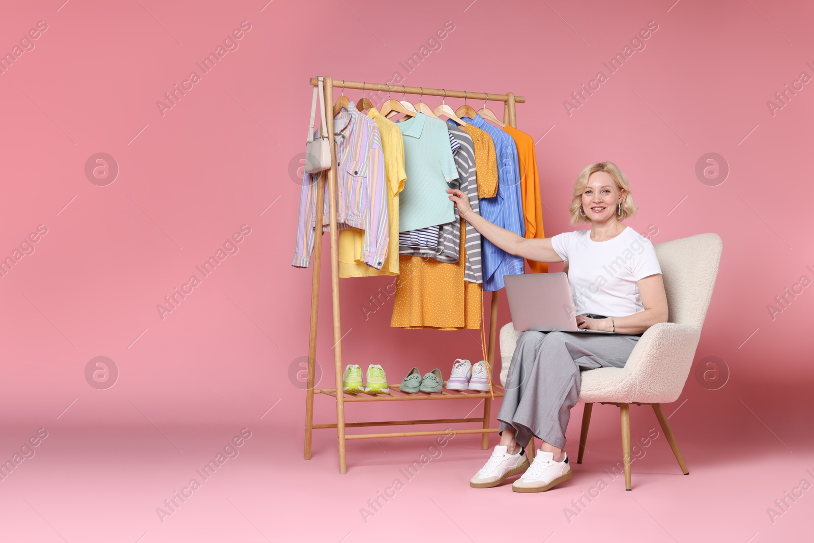 Smiling fashion stylist with laptop and clothing rack on pink background. Space for text Photo of Smiling fashion stylist with laptop and clothing rack on pink background. Space for text