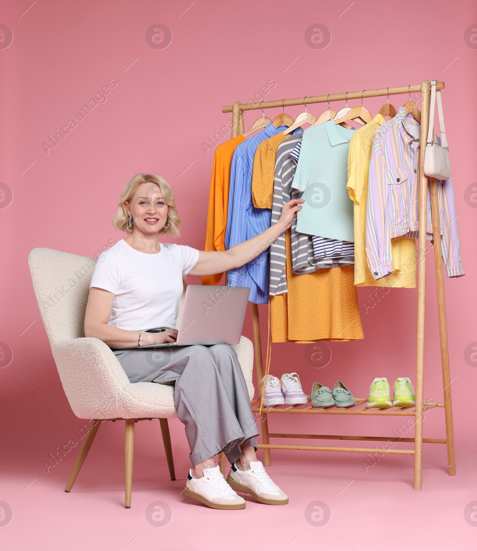 Smiling fashion stylist with laptop and clothing rack on pink background Photo of Smiling fashion stylist with laptop and clothing rack on pink background