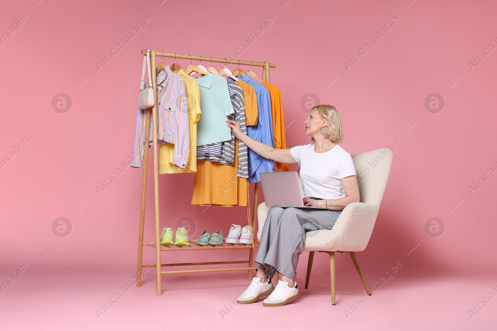 Smiling fashion stylist with laptop and clothing rack on pink background Photo of Smiling fashion stylist with laptop and clothing rack on pink background