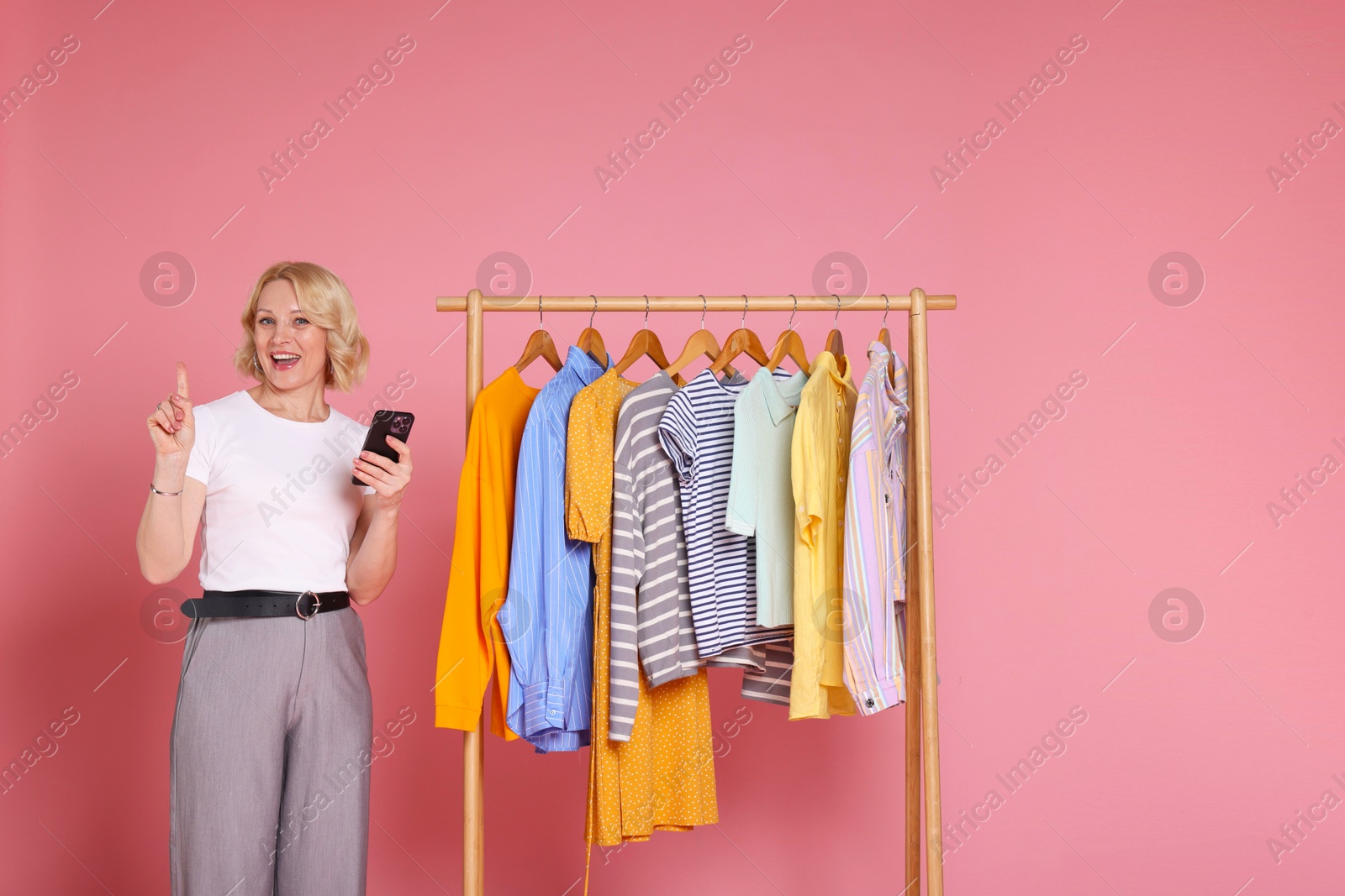 Happy fashion stylist with smartphone and clothing rack on pink background Photo of Happy fashion stylist with smartphone and clothing rack on pink background