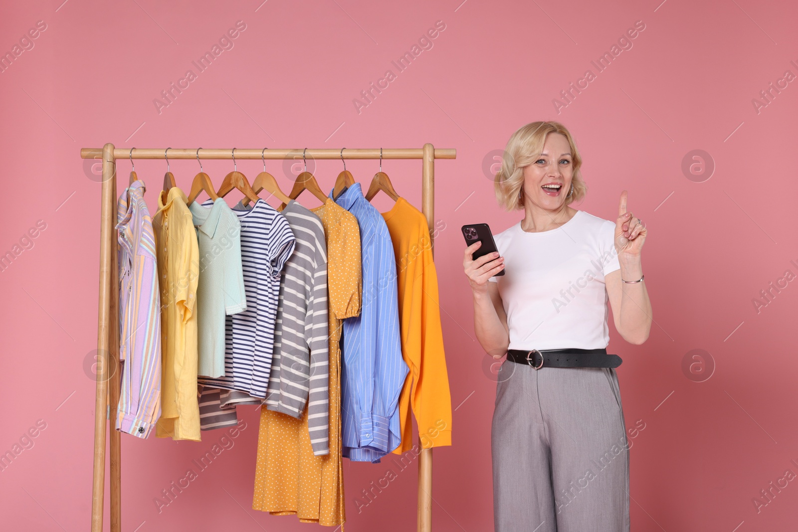 Happy fashion stylist with smartphone and clothing rack on pink background Photo of Happy fashion stylist with smartphone and clothing rack on pink background