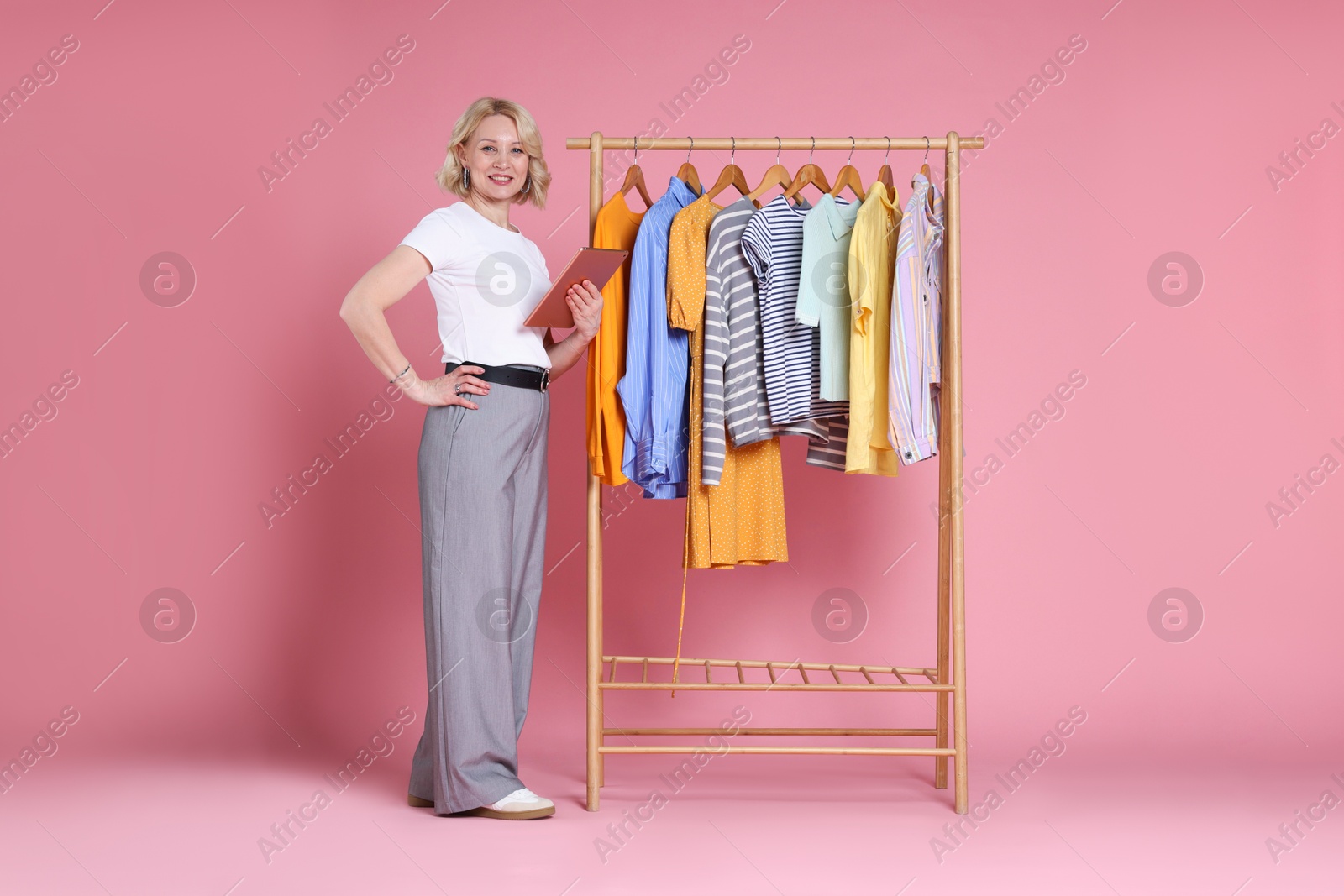 Smiling fashion stylist with tablet and clothing rack on pink background Photo of Smiling fashion stylist with tablet and clothing rack on pink background