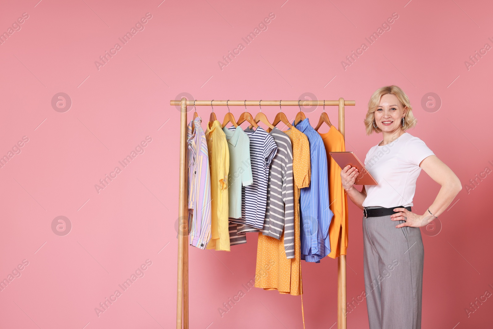 Smiling fashion stylist with tablet and clothing rack on pink background. Space for text Photo of Smiling fashion stylist with tablet and clothing rack on pink background. Space for text