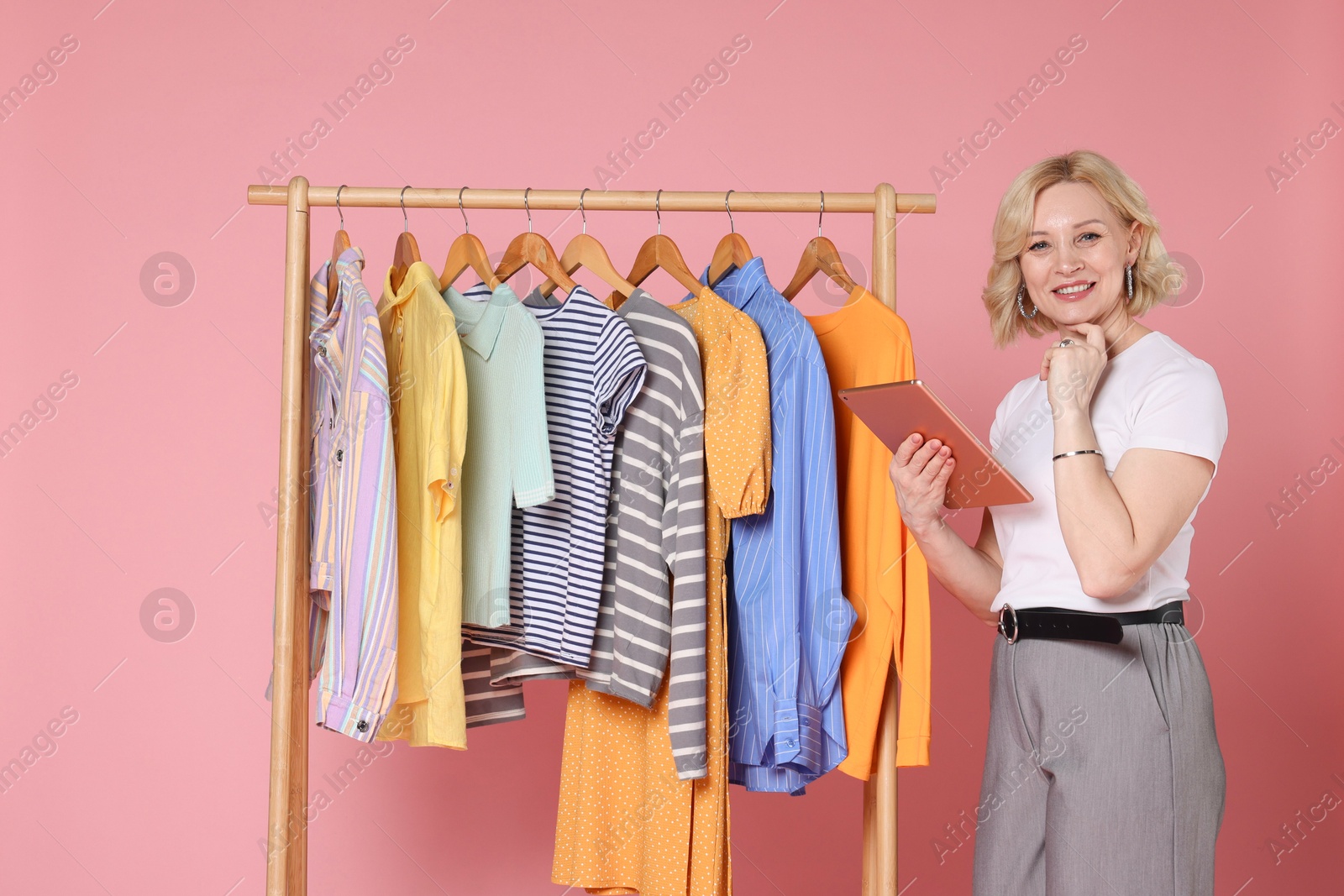 Smiling fashion stylist with tablet and clothing rack on pink background Photo of Smiling fashion stylist with tablet and clothing rack on pink background