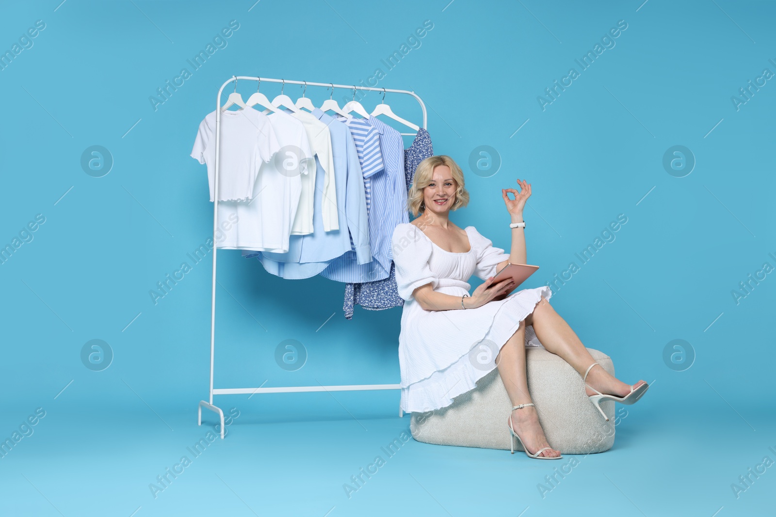 Smiling fashion stylist with tablet showing ok gesture and clothing rack on light blue background Photo of Smiling fashion stylist with tablet showing ok gesture and clothing rack on light blue background