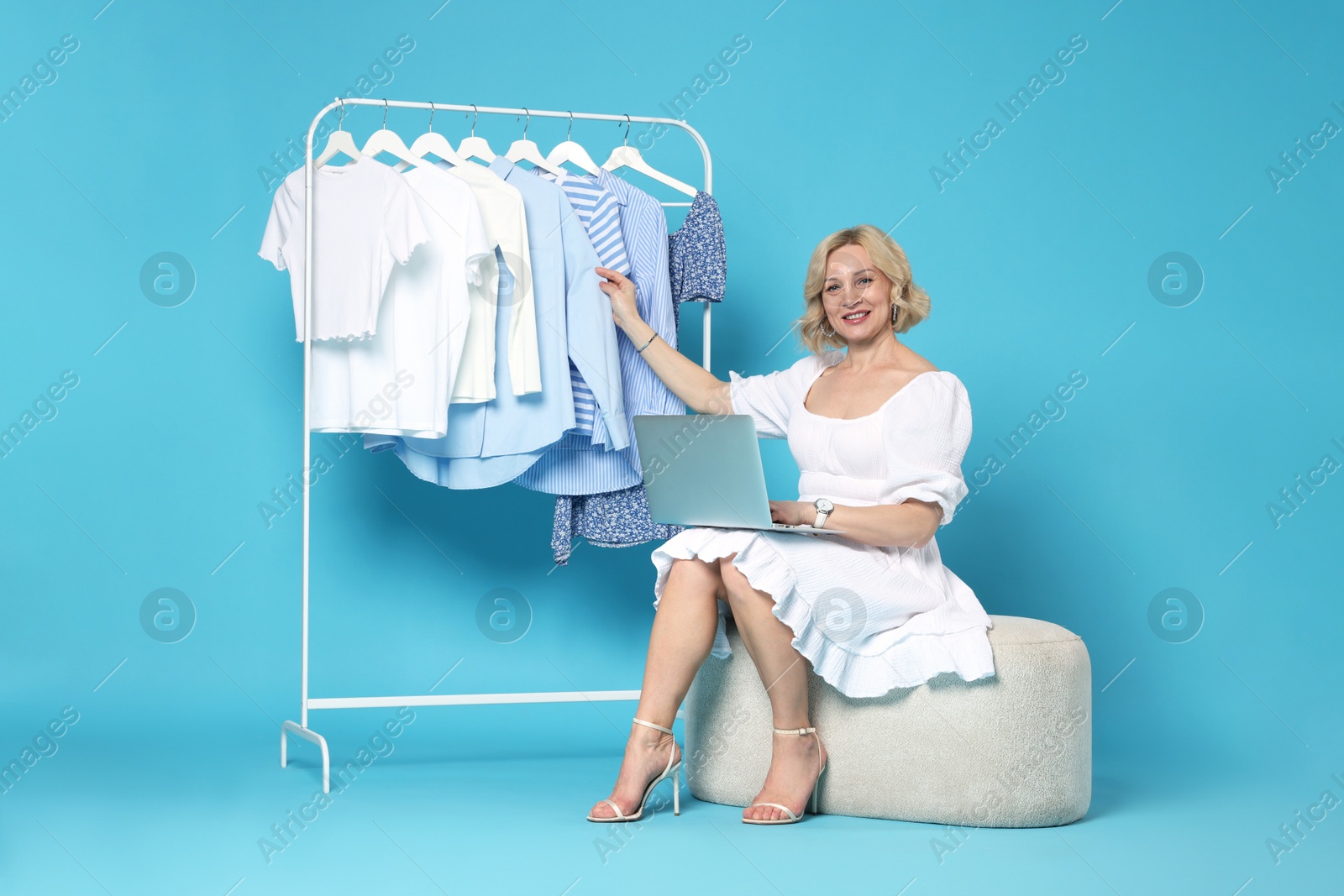 Smiling fashion stylist with laptop and clothing rack on light blue background Photo of Smiling fashion stylist with laptop and clothing rack on light blue background