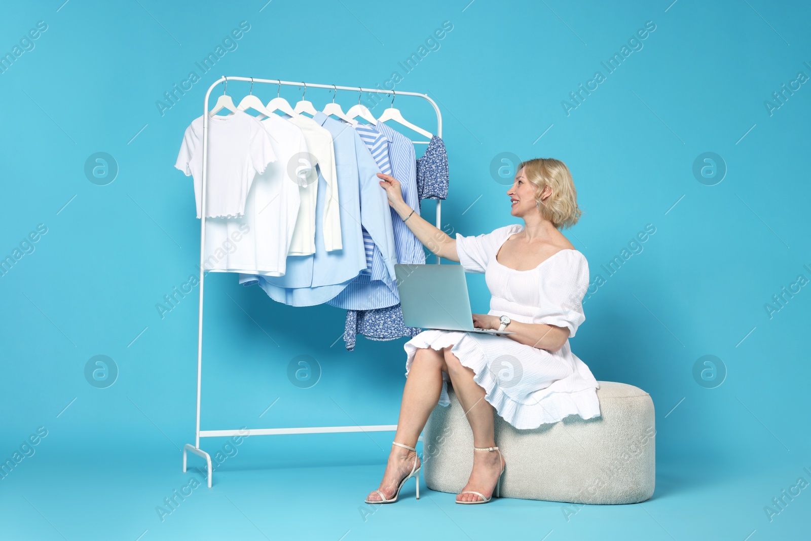 Smiling fashion stylist with laptop and clothing rack on light blue background Photo of Smiling fashion stylist with laptop and clothing rack on light blue background
