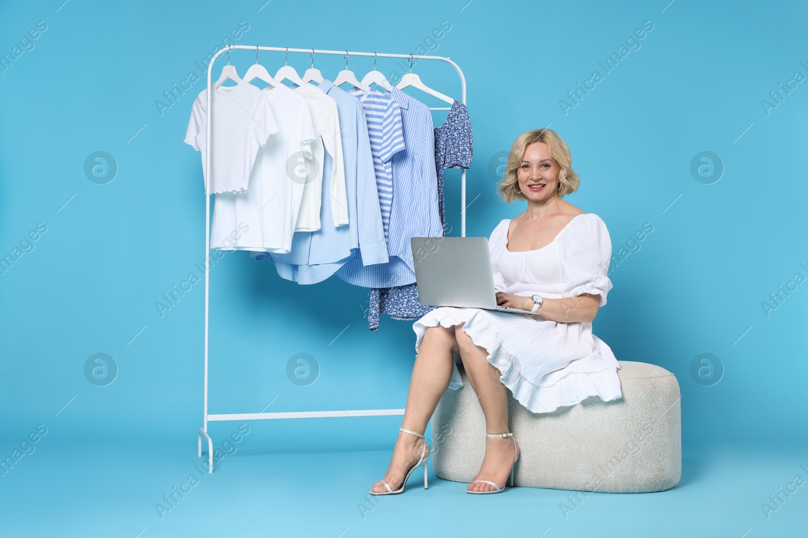 Smiling fashion stylist with laptop and clothing rack on light blue background Photo of Smiling fashion stylist with laptop and clothing rack on light blue background