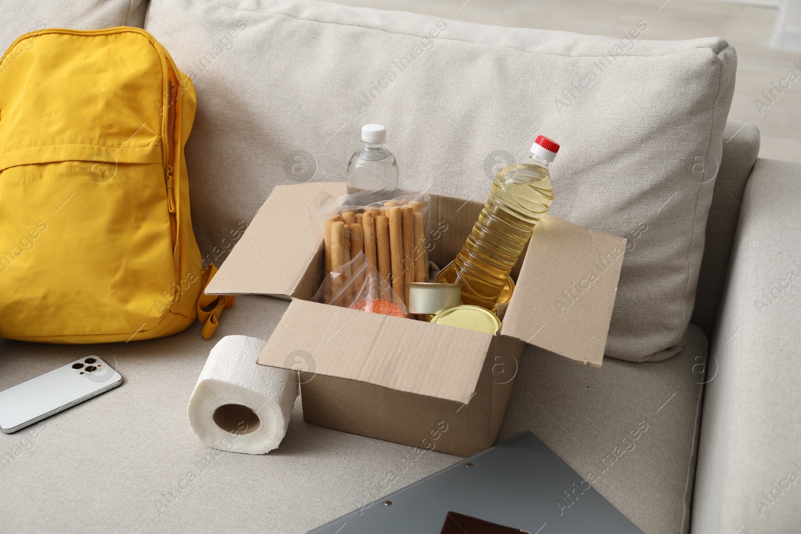 Survival kit. Different emergency supplies and food products in box on sofa Photo of Survival kit. Different emergency supplies and food products in box on sofa