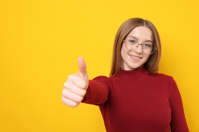 Photo of Portrait of smiling teenage girl showing thumbs up on yellow background. Space for text