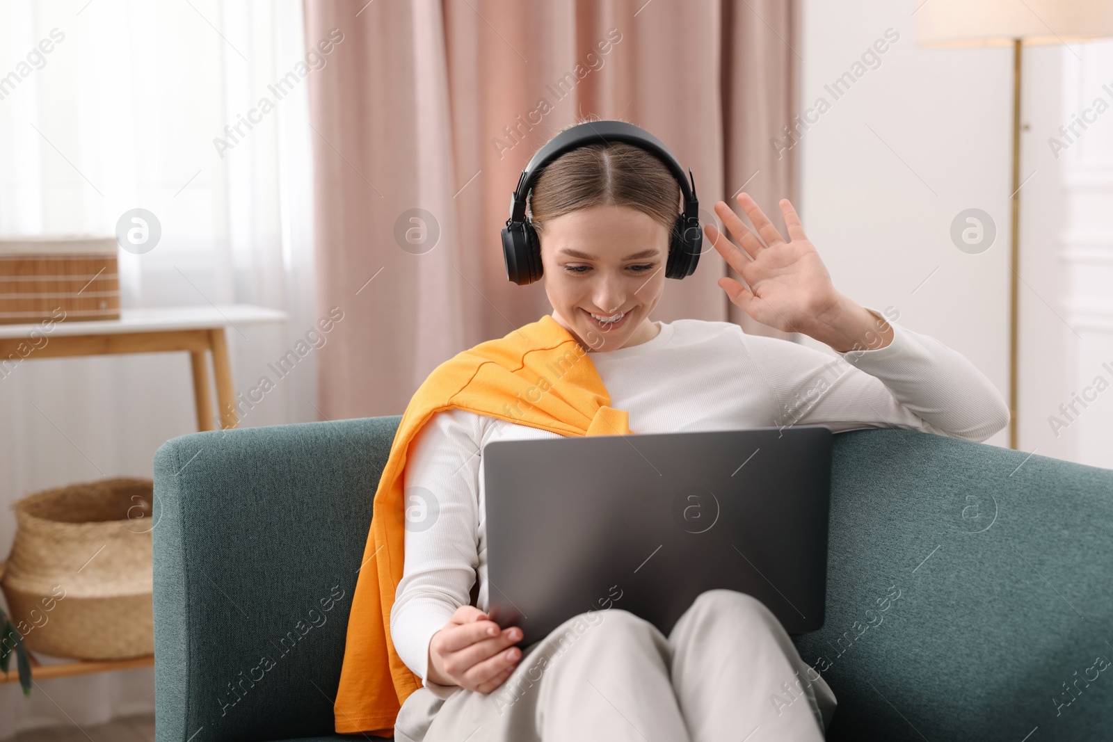 Photo of Smiling teenage girl in headphones having videochat by laptop on sofa at home