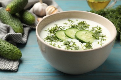 Photo of Tasty cucumber soup in bowl and ingredients on light blue wooden table, closeup