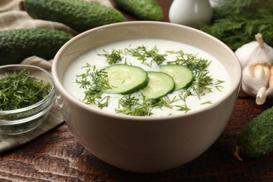 Photo of Tasty cucumber soup in bowl and ingredients on wooden table, closeup