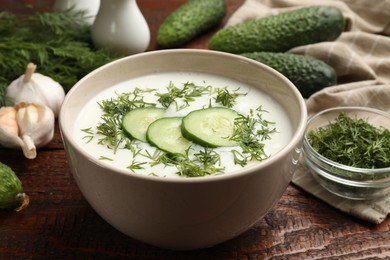 Photo of Tasty cucumber soup in bowl and ingredients on wooden table, closeup
