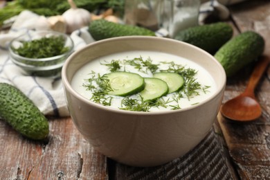 Photo of Tasty cucumber soup in bowl and ingredients on wooden table, closeup
