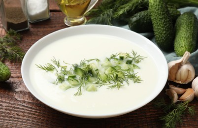 Photo of Tasty cucumber soup in bowl and ingredients on wooden table, closeup