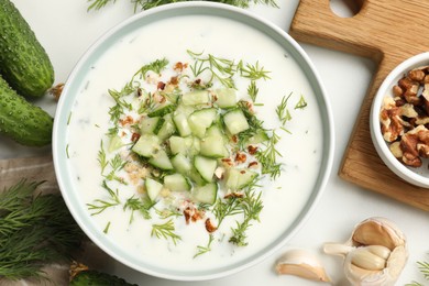 Photo of Tasty cucumber soup with dill and nuts in bowl among products on white table, flat lay