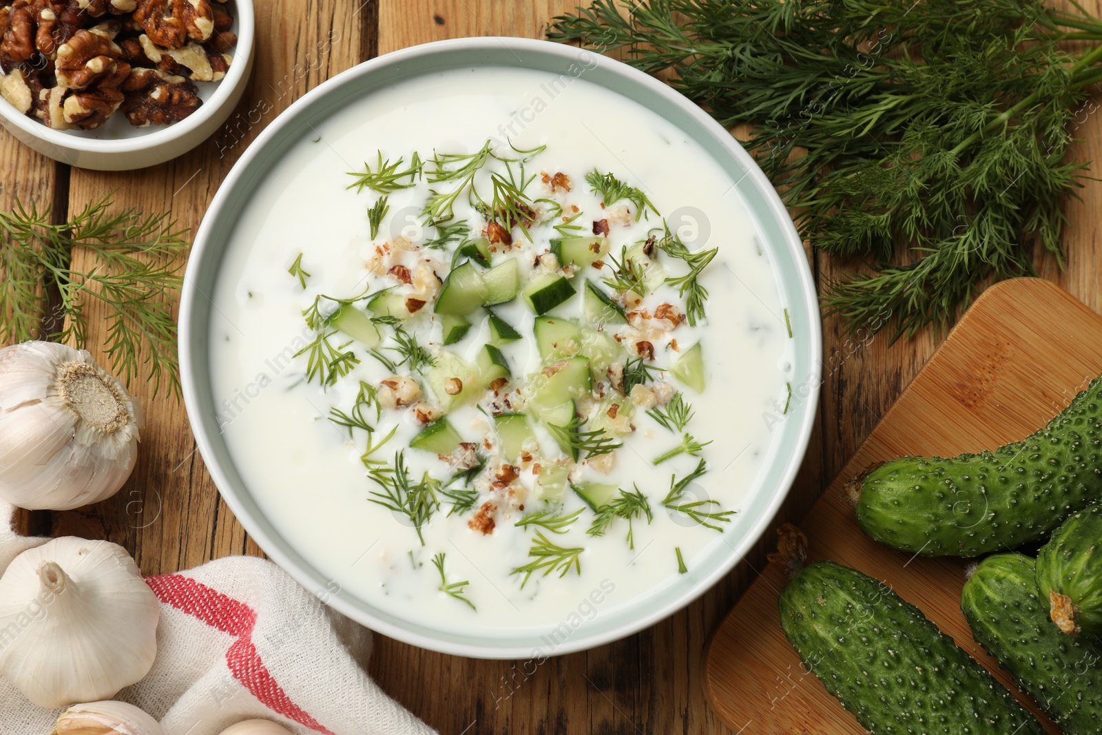 Tasty cucumber soup with dill and nuts in bowl among products on wooden table, flat lay Photo of Tasty cucumber soup with dill and nuts in bowl among products on wooden table, flat lay